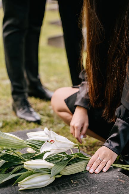A person with long brown hair is kneeling at a grave, arranging a bouquet of fresh white lilies with large green leaves placed on the headstone. The individual is wearing a dark coat, and their hands 