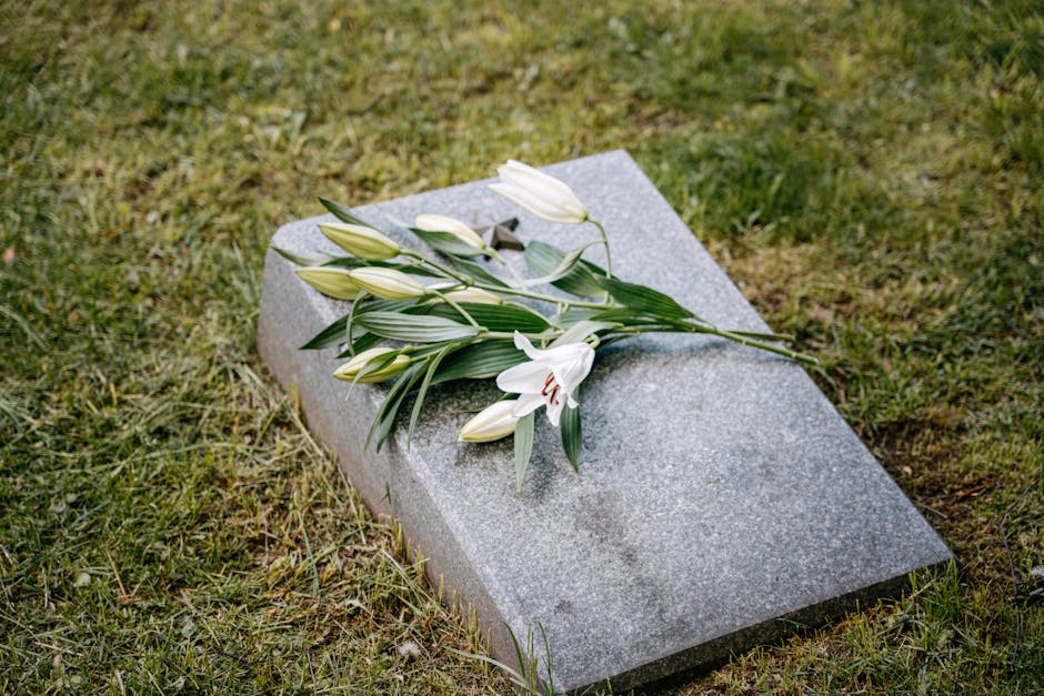 A bouquet of white lilies with closed buds and long green leaves rests on a light gray granite memorial plaque lying flat on a patch of grass. The lilies' petals are smooth and fresh, with some flower