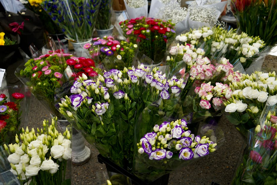 A vibrant display of fresh flowers arranged in bouquets and wrapping in a florist shop. The arrangements include purple and white lisianthus, white roses, pink and white spray roses, and small daisy-l